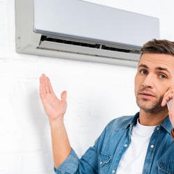 Man standing next to a ductless AC unit while on the phone with his other hand up in the air